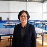 东莞市光明小学校长陈芳的正面照片，她戴着眼睛，站在学校新落成的体育馆内，身后是整齐的乒乓球训练区